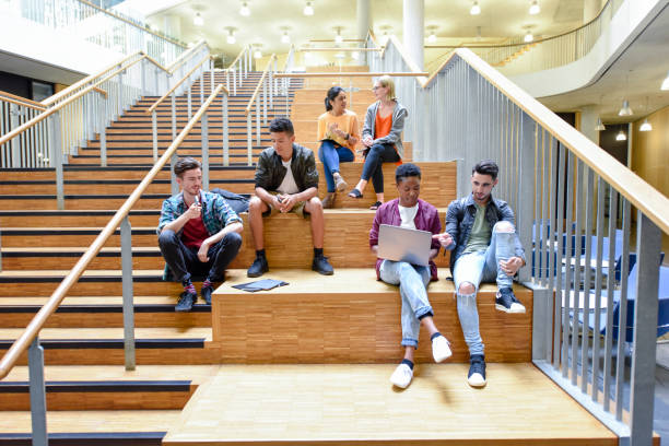College friends sitting on steps in modern interior, chatting and using laptop