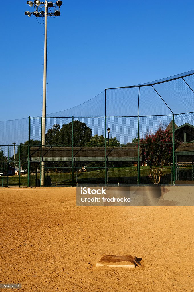 Baseball Field at a Baseball Game this photo is of a baseball infield of a baseball field at a baseball game. the photo includes third base, home plate and the baseball player's dugout. the baseball field is located at a local park or highschool. there is also stadium lights in the background. and there are also trees and grass in the background. the picture was taken during the spring or summer with clear blue sky in the background. the lighting is natural sunlight.  Baseball - Ball Stock Photo