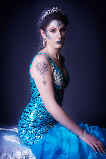 Vertical colour studio shot on dark gray of young woman. Three quarter length, seated on bench covered in white satin. Side view looking at camera with serious expression. Makeup with gems. Hair sprayed with sparkly silver and braided on top with tiara.