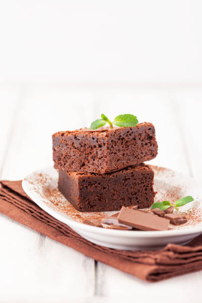 istock Chocolate brownie square pieces in stack on white plate decorated with mint leaves and cocoa powder on white vintage wooden background. American traditional delicious dessert. Close up photography 914330808