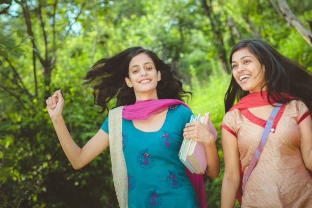 Outdoor low angle image of female Indian college students walking together on the way from their village to college surrounded by lush green trees and foliage. They are in their traditional dress of north India (salwar kameez and dupatta). Two people, selective focus with copy space. Shoot location Solan, (a city in mountain) Himachal Pradesh, India.