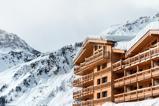 Taken from street with several common buildings. Color photography of multiple wooden common chalet in ski resort in front of beautiful snowcapped Alps mountain peak with fresh white snow and overcast sky with many clouds. This image was taken in winter season in Val d'Isere, a french ski resort village in Tarentaise mountains, in Savoie, Auvergne-Rhone-Alpes region in European Alps.
