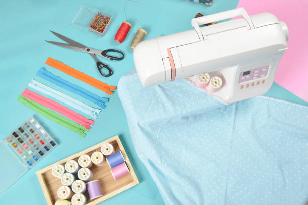 istock Fabrics on sewing machine amid the scissors, shirt buttons, zipper, pin and colorful thread rolls for sewing on blue background, Sewing and needlework concept. 914457406