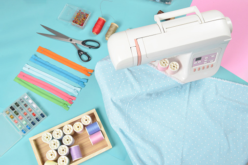 Fabrics on sewing machine amid the scissors, shirt buttons, zipper, pin and colorful thread rolls for sewing on blue background, Sewing and needlework concept.