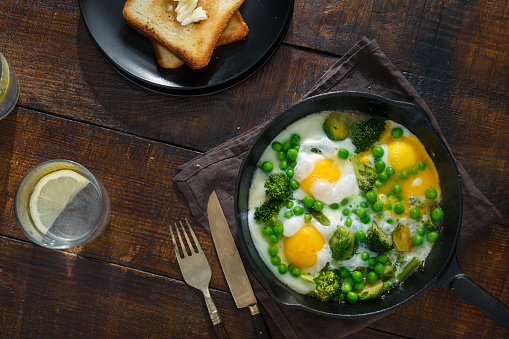 Full breakfast table with fried eggs with brussels sprouts, beans, broccoli and green peas in frying pan on dark wooden table with toasts and soda with lemon, top view