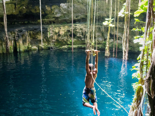 Young man rope swinging into Cenote sink hole Oxman Young man using a rope swing to jumpp into Cenote sink hole Oxman in Yucatan, Mexico, 2017 cenote stock pictures, royalty-free photos & images