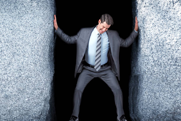 businessman attempting to widen opening between two large rocks - claustrophobic imagens e fotografias de stock