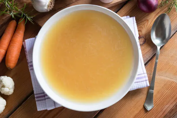 Bone broth made from chicken on a wooden table, with vegetables in the background, top view