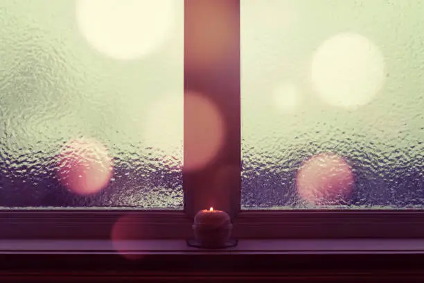 Winter evening. Frosted window, burning candle and purple bokeh lights.