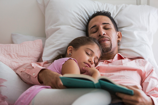 A father reading a bedtime story to his 7 year old daughter. The little girl is resting her head on daddy's shoulder and they both have fallen asleep. They are mixed race Hispanic, Caucasian and black.