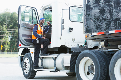 A semi-truck driver climbing into the truck cab, smiling at the camera. The mature black man in his 40s is wearing a reflective vest.