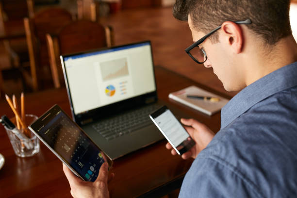 istock Attractive man in glasses working with multiple electronic internet devices. Freelancer businessman has laptop and smartphone in hands and laptop on table with charts on screen. Multitasking theme 914622582