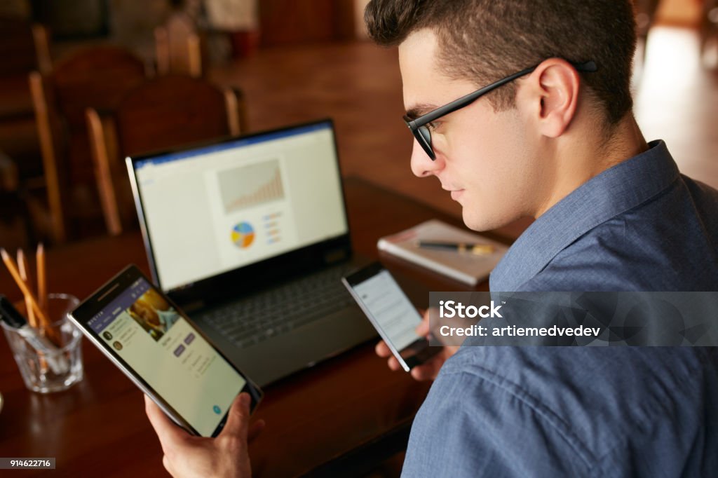 Hombre atractivo en vasos con múltiples dispositivos electrónicos de internet. Empresario autónomo tiene laptop y smartphone en las manos y laptop en mesa de cartas en pantalla. Tema de la multitarea - Foto de stock de Variación libre de derechos