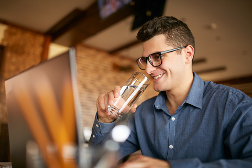 Handsome caucasian man with glass of water in hand works with laptop. Businessman in glasses drinks water for body hydration while working. Attractive designer quench thirst.