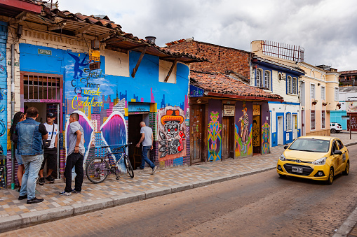 Bogota, Colombia - May 28, 2018: Some local Colombians are seen chatting on the sidewalk of a colorful street in the historic La Candelaria District in the capital city of Bogota in the South American country of Colombia. Many of the walls in the area are painted in the vibrant colours of Colombia, sometimes depicting Pre-Colombian legends or modern Street Art and Graffiti. A Taxi drives past. Photo shot in the late afternoon sunlight in the Golden Hour; horizontal format. Camera: Canon EOS 5D MII. Lens: Canon EF 24-70mm F2.8L USM.