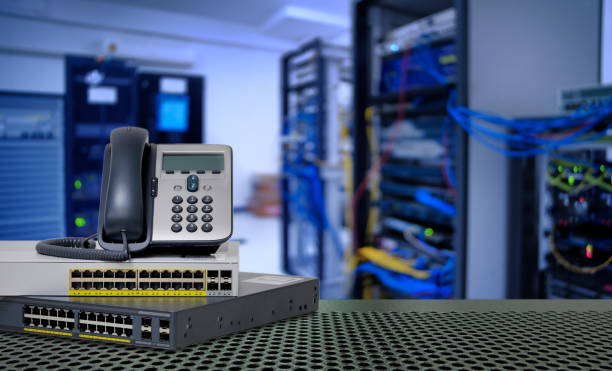 A desktop VoIP phone rests on top of a network switch, with a blurred background featuring server racks and colorful cables in a technology environment.