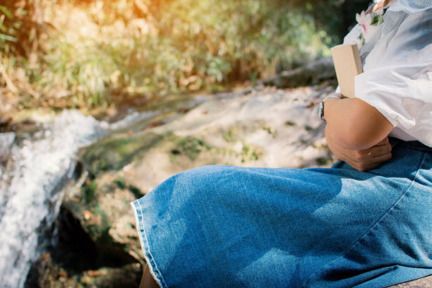 istock Midsection Of Woman Holding Book While Sitting On Rock By Waterfall 914707554