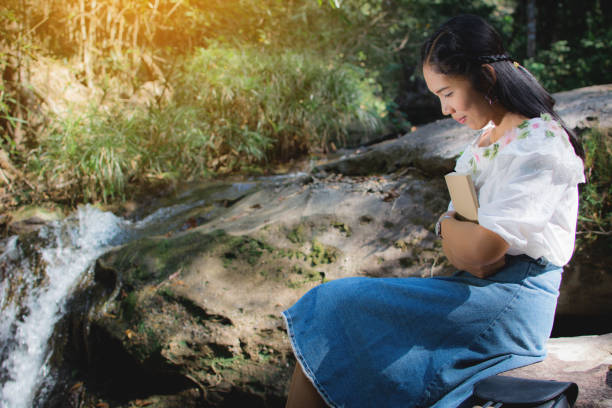 istock Young Woman Holding Book While Sitting On Rock By Waterfall 914708142