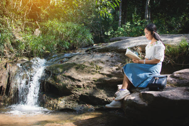 istock Young Woman Reading Book While Sitting On Rock By Waterfall 914714930