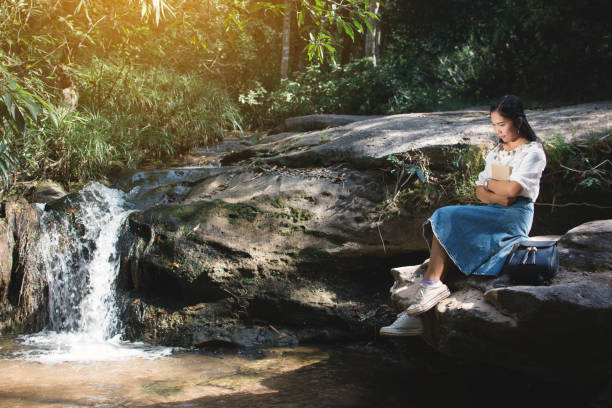 istock Young Woman Holding Book While Sitting On Rock By Waterfall 914717960
