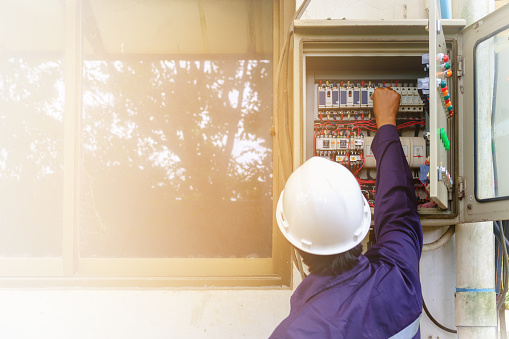 Back of asian engineer or technician in safety uniform and white security helmet turning on switch in the electrical cabinet with copy space and sun light effect for industrial concept