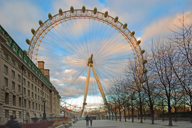 The Millennium Wheel isa popular tourist attraction on the River Thames in London, and affords panoramic views over the skyline of London, England, 2018 The London Eye on the South Bank taken early in the morning with two people walking.  It is a popular tourist attraction and you can see panoramic views over the City of London,  Great Britain, February 2018 london county hall stock pictures, royalty-free photos & images