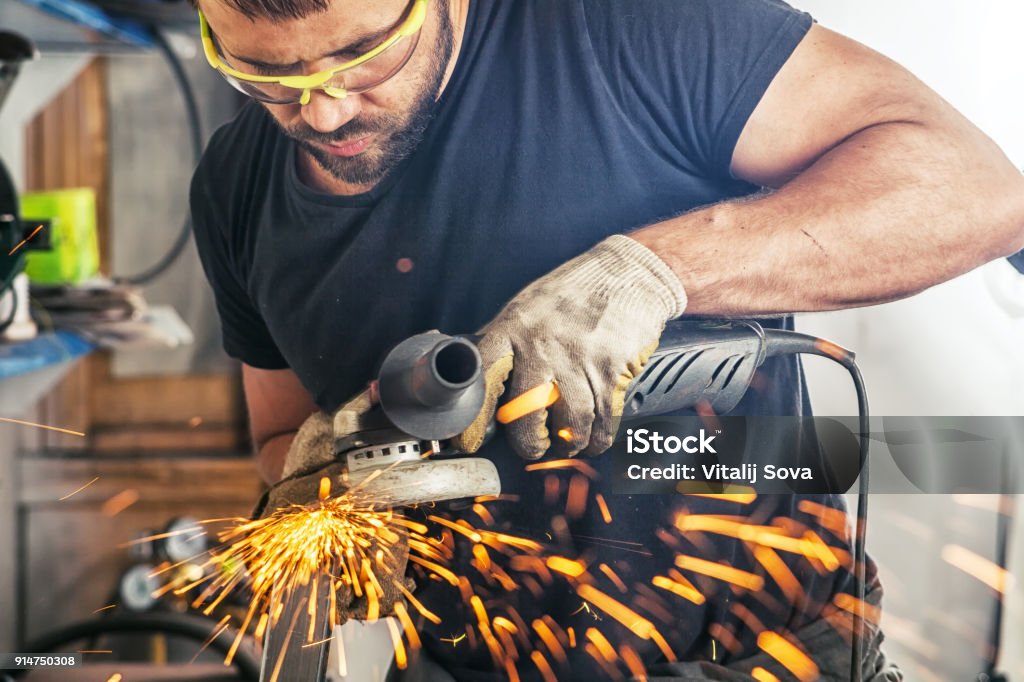 man processes metal an angle grinder A young man welder in a black T-shirt, goggles and construction gloves processes metal an angle grinder   in the garage, in the background a lot of tools, sparks fly to the side Adult Stock Photo