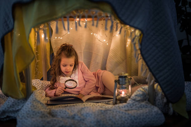 istock Small girl using magnifying glass while reading a book in a tent. 914750706