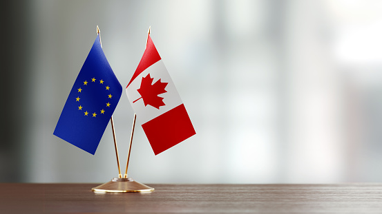 European Union and Canadian flag pair on desk over defocused background. Horizontal composition with copy space and selective focus.