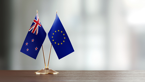 European Union and  New Zealand flag pair on desk over defocused background. Horizontal composition with copy space and selective focus.