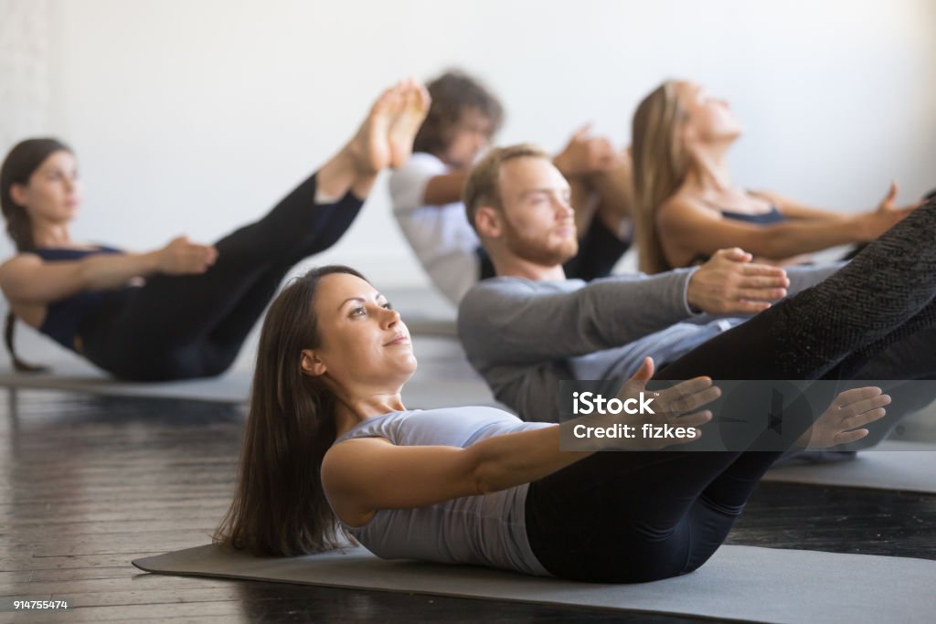 Group of young sporty people in boat pose Group of young sporty people practicing fitness lesson with instructor, doing warming up exercise, boat pose, working out, students training in club, friends at fitness lesson, indoor, studio Pilates Stock Photo