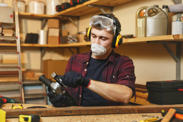 Caucasian young man in plaid shirt, black T-shirt, noise insulated headphones, protective mask working in carpentry workshop at wooden table place with different tools, sawing iron with power saw joven caucásica en auriculares de aislamiento de ruido de camisa a cuadros, camiseta negra, máscara protectora trabajando en taller de carpintería en el lugar de la mesa de madera con diferentes herramientas, cortar hierro con poder vio.