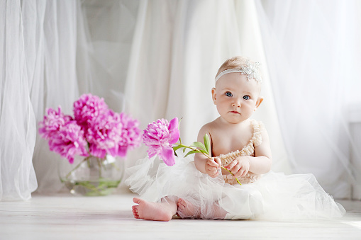 Little pretty girl with blue eyes sits on a floor with flowers of a peony