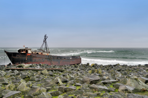 A shipwreck on Saint Paul Island in the Bering sea. The beach is a conglomeration of jagged rocks interspersed with green algae. Looking closely at the bow, there is a Northern Fur Seal resting on the rocks.