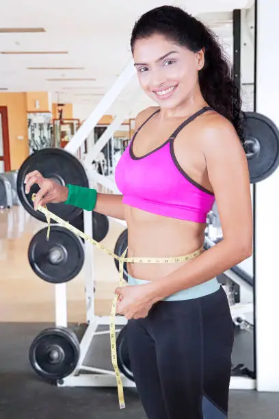 Portrait of African woman smiling at the camera while measuring her slim waist with a measure tape in the gym center
