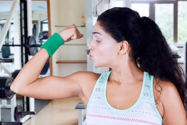 Image of Indian woman looks proud while showing her arm muscle and standing in the fitness center