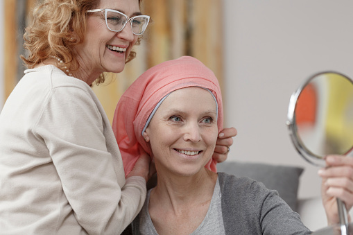 Happy woman with cancer in pink headscarf looking in the mirror while mother smiling
