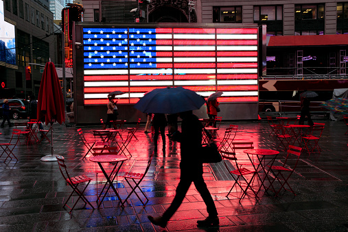City in the Rain: pedestrian carrying umbrella and shoulder bag silhouette against the famous illuminated American Flag in very wet, dark Times Square