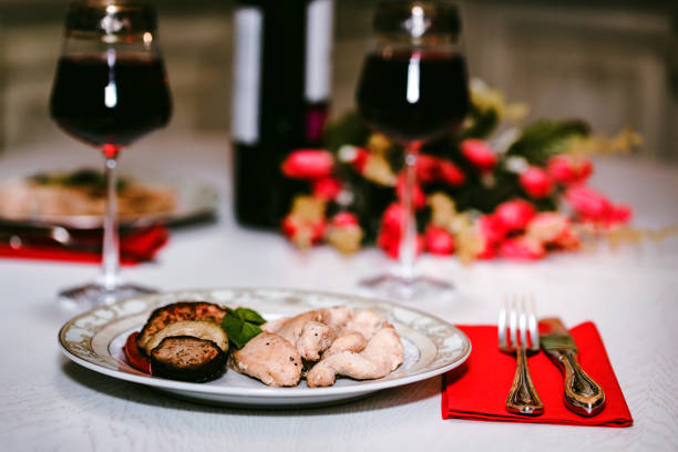 Close up table served for romantic dinner for two with glasses of red wine, roasted chicken fillet with vegetables on plate and flowers. St.Valentine's concept
