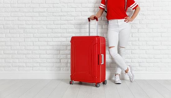 concept travel and tourism. legs of a girl with a red suitcase near  white empty brick wall