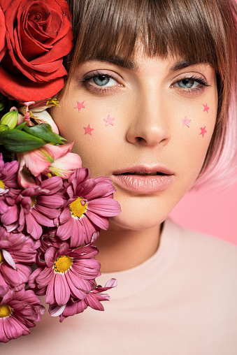 Young woman with stars on her face posing with bright flowers near her face and looking at camera