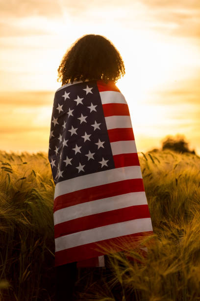 vintage style photograph of mixed race african american girl teenager female young woman in a field of wheat or barley crops wrapped in usa stars and stripes flag in golden sunset evening sunshine - depression sadness usa american flag imagens e fotografias de stock