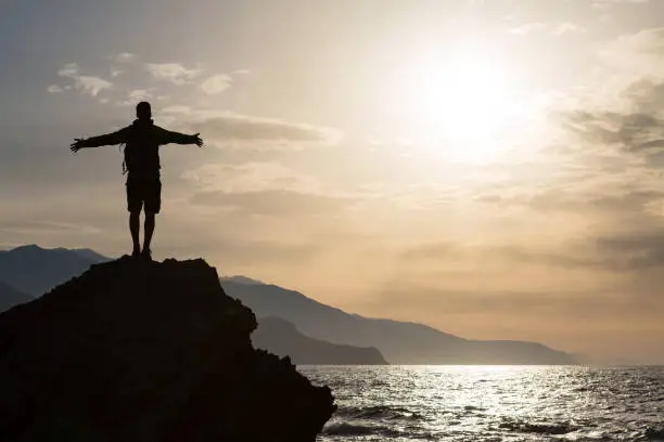 Man with arms outstretched celebrating or praying in beautiful inspiring sunrise with mountains and sea. Man hiking or climbing with hands up enjoy inspirational landscape on rocky top on Crete.