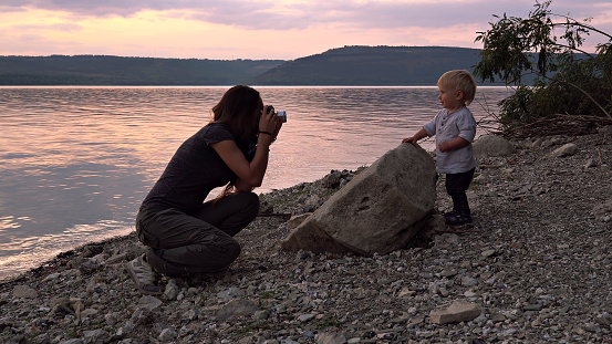 Woman with retro camera taking photos of her little son on shore of lake at sunset. Concept of happy family relationship