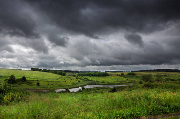 Cloudy summer landscape with small river.Dark stormy clouds in dramatic overcast sky.Fields,green meadows and woods.