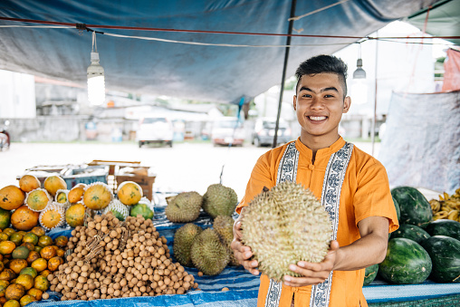 Portrait of young farmer or green market salesman, who is selling fresh fruits and vegetables on the market in small town in South East Asia. Cheerful young man is making money with selling his home grown farm products
