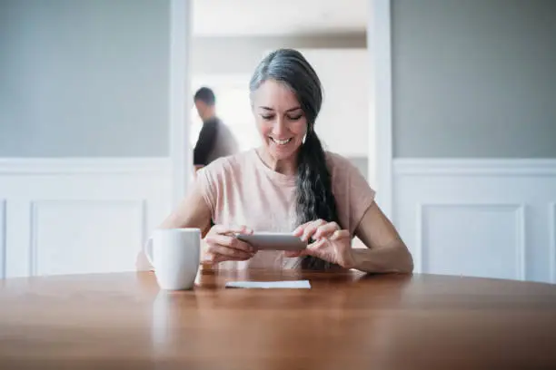 A woman in her 50's in her home takes a picture of a check for remote deposit capture to her bank account.  Depicts the ease of online banking with smart phones.