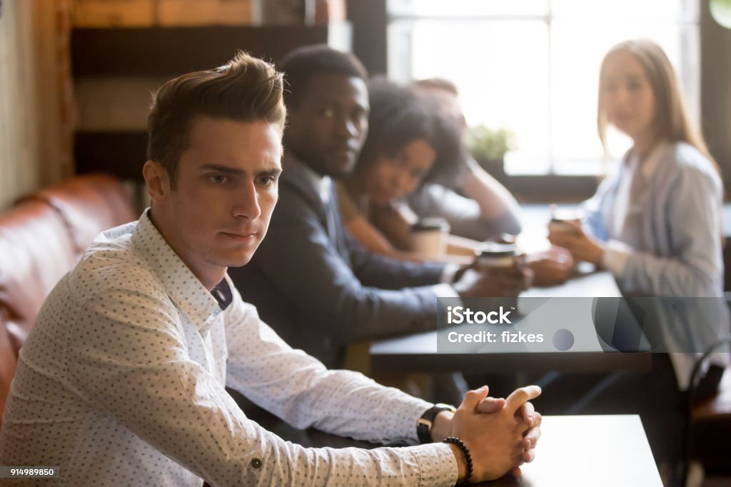 Diverse friends looking at frustrated man sitting alone in cafe Diverse people looking at thoughtful frustrated man in cafe, sad misunderstood guy feels offended ignoring multiracial friends sitting alone at table, excluded outstand person suffers from bullying Group Of People Stock Photo