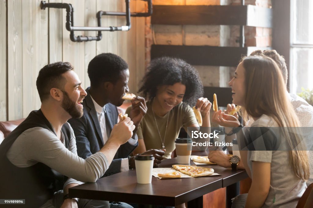 Multiracial happy young people laughing eating pizza together in pizzeria Multiracial happy young people eating pizza in pizzeria, black and white cheerful mates laughing enjoying meal having fun sitting together at restaurant table, diverse friends share lunch at meeting Friendship Stock Photo