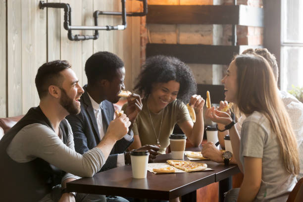 multiracial happy young people laughing eating pizza together in pizzeria - grupo pequeno de pessoas imagens e fotografias de stock